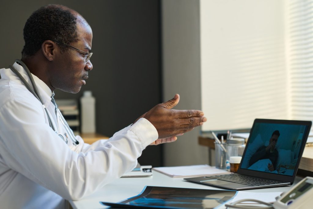 Confident mature teledoc sitting in front of laptop with sick patient on screen